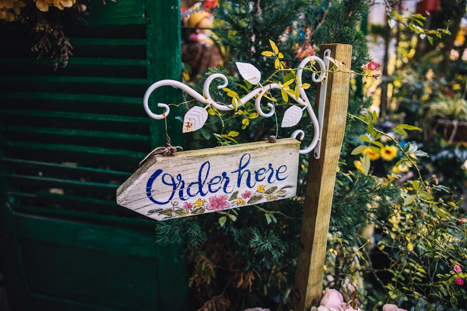 A rustic wooden sign with the handwritten word 'Order here' in blue lettering, hanging from a decorative white wrought iron bracket attached to a wooden post, amidst lush green foliage and blooming fl