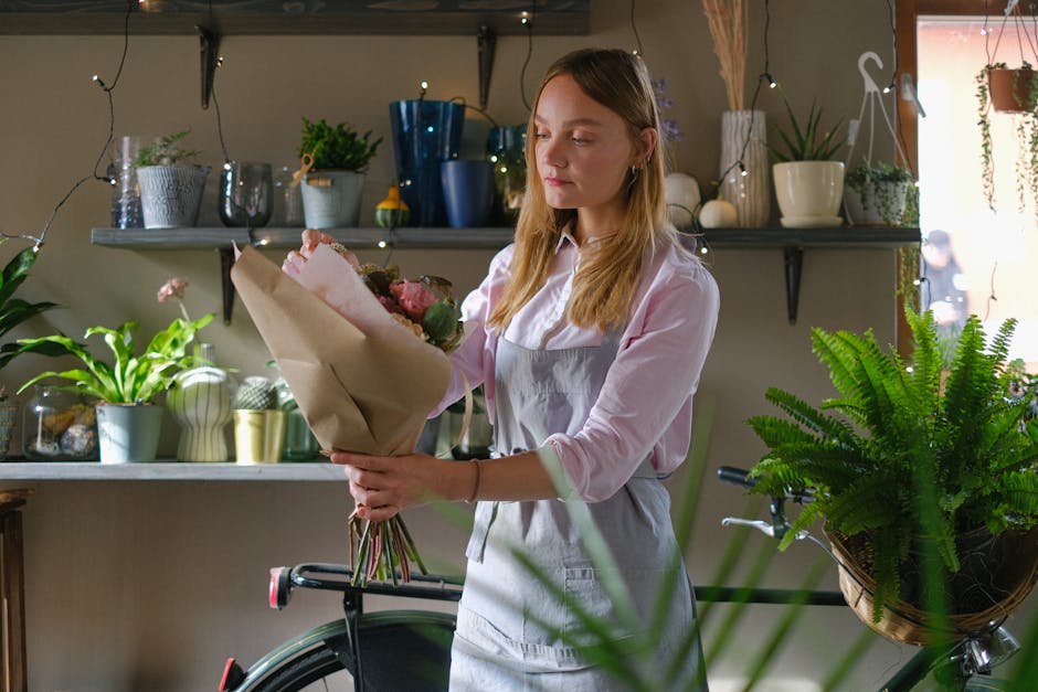 A young woman with long, light brown hair and fair skin, wearing a pink shirt and a light gray apron, is holding a large bouquet of fresh flowers wrapped in brown paper inside a cozy flower shop. The 