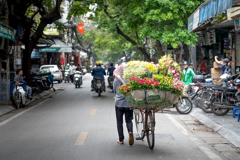 A street scene featuring a person pushing a bicycle loaded with a large, colorful floral arrangement. The bouquet includes pink roses, white hydrangeas, yellow tulips, and green foliage, all arranged 