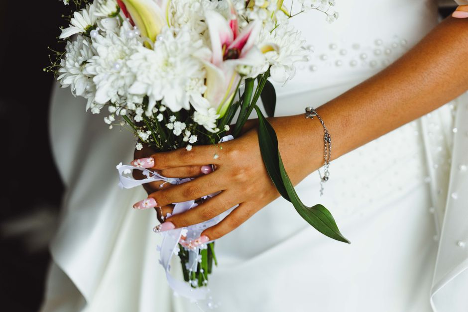 A close-up of a bride's hand holding a bridal bouquet composed of white lilies, chrysanthemums, and baby's breath, with accents of pink flowers. The bouquet is wrapped in white satin ribbon with pearl