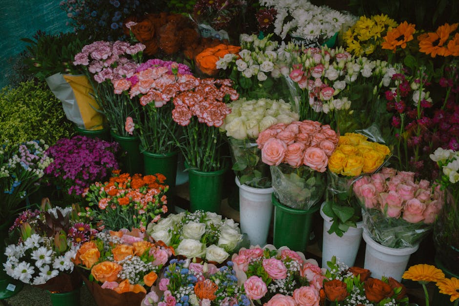 A vibrant display of fresh flowers arranged in various bouquets and potted plants. The collection includes pink, white, yellow, orange, and purple blossoms such as roses, carnations, and daisies, with